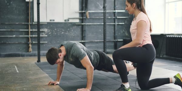 Strong guy is doing push-ups in sports gym while trainer young woman is speaking motivating student. Healthy activity, lifestyle and sports concept.